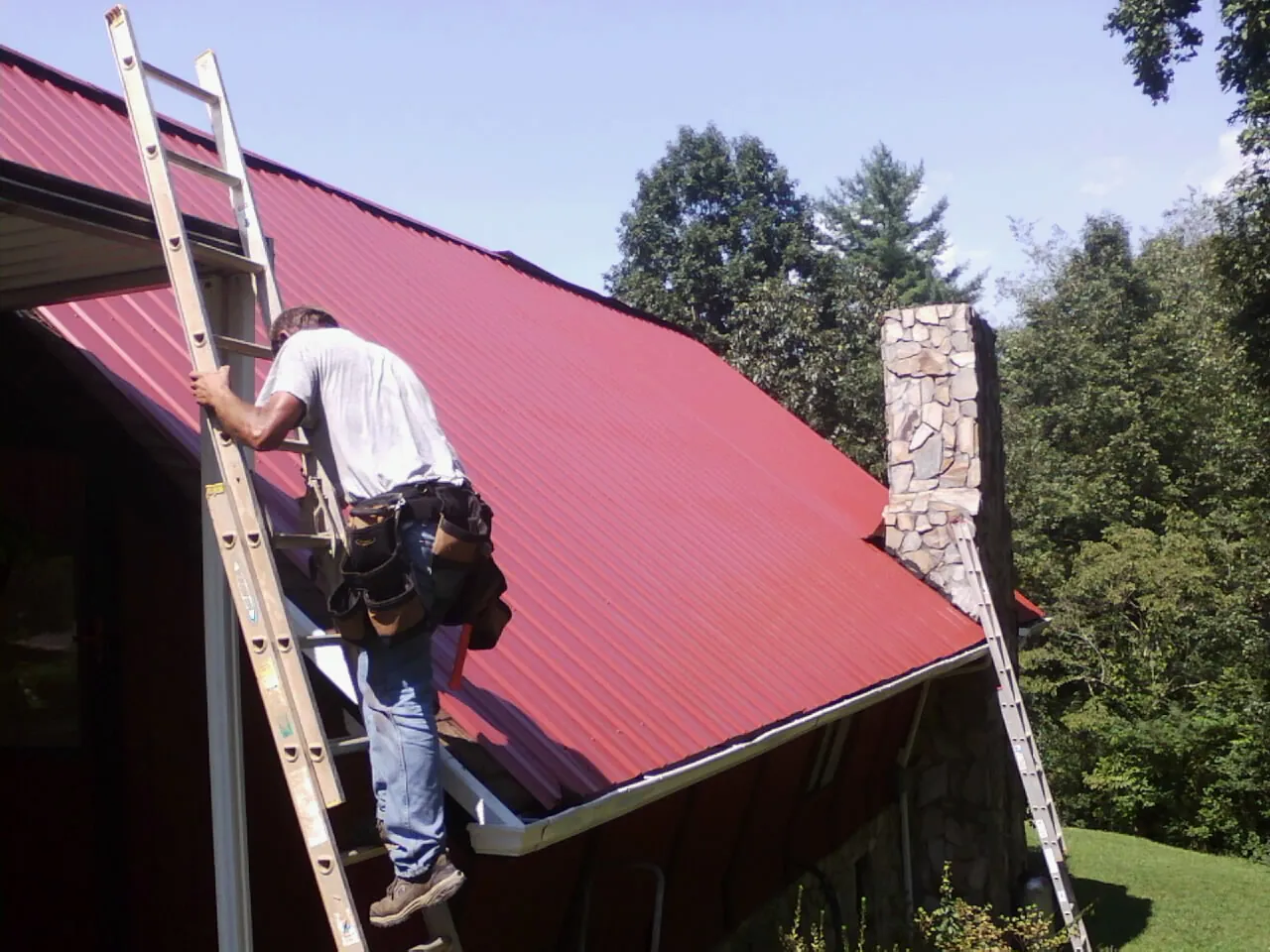 Roofer on a ladder beside a red metal roof and stone chimney — Soffit & Fascia Repair by Roofing Brothers in Peacham