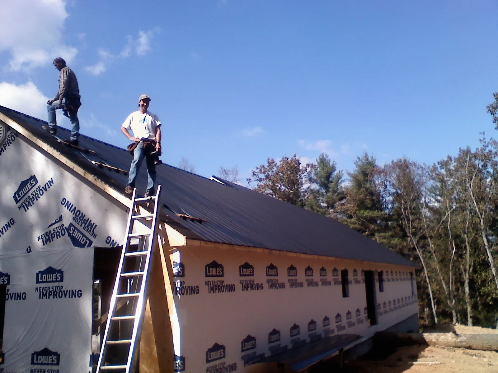 Two roofers standing on a long dark metal roof over a wrapped building in Peacham